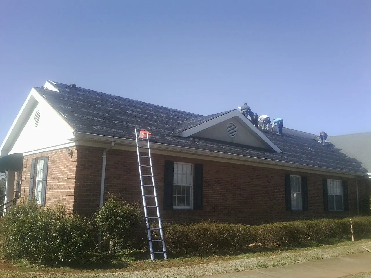 Crew tearing shingles off a brick ranch roof with a ladder set — Emergency Roof Repair work by our crew in Deadwood